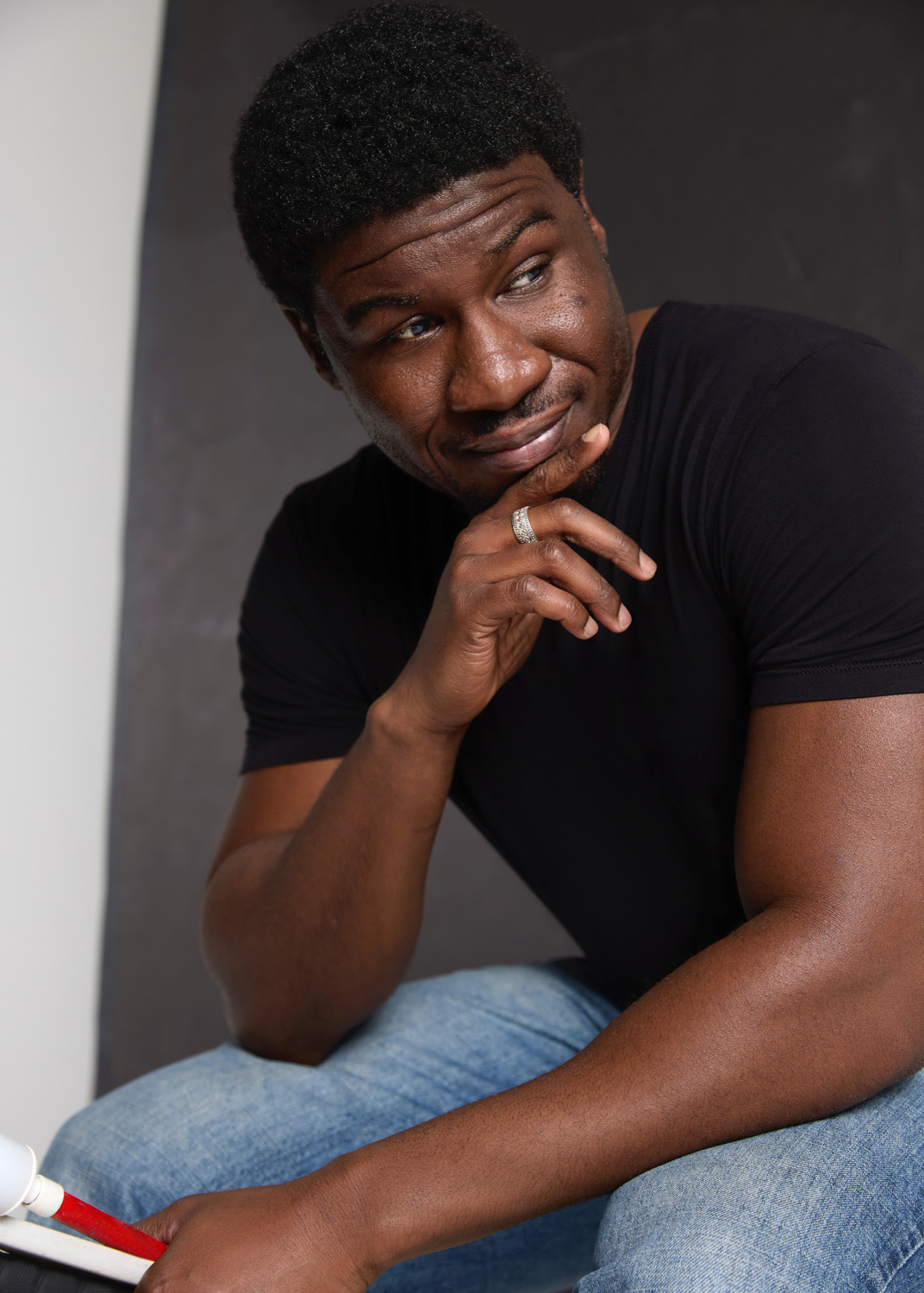 Ben Akuoko, a man with dark skin wearing a black t-shirt and jeans, sits contemplatively with his white cane in one hand
