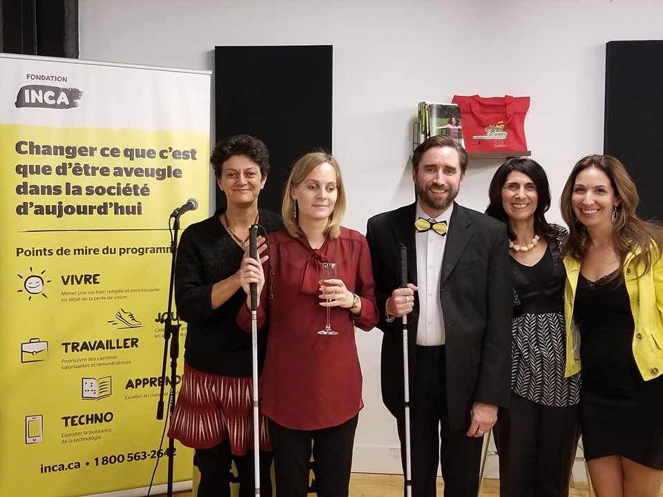 Nima Machouf, Catheryne, David, Valérie et Sarah posent devant la bannière et le podium d'INCA. Catheryne et David tiennent leur canne blanche. David et Sarah sont assortis aux couleurs jaune et noir d'INCA.