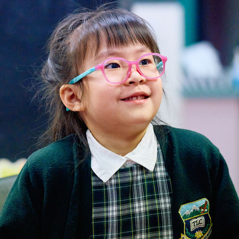 A smiling young child with dark hair and medium skin wearing bright pink-and-turquoise glasses and a green plaid school uniform.