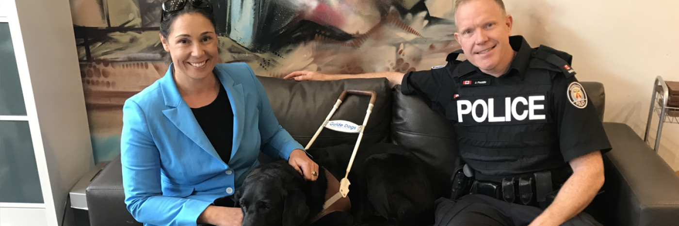 Victoria, her Guide Dog Alan, and a police officer sit on a couch.