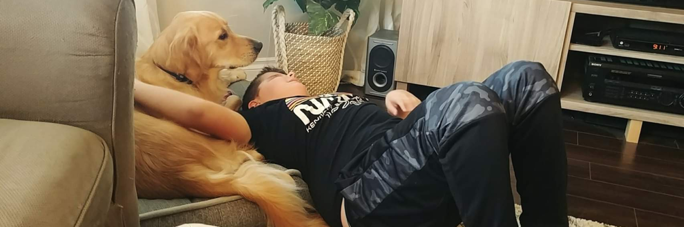 Landon and his CNIB Buddy Dog Ruggles, a Golden Retriever, laying on a cushion together in their living room. Landon’s arm is around Ruggles, petting his back.