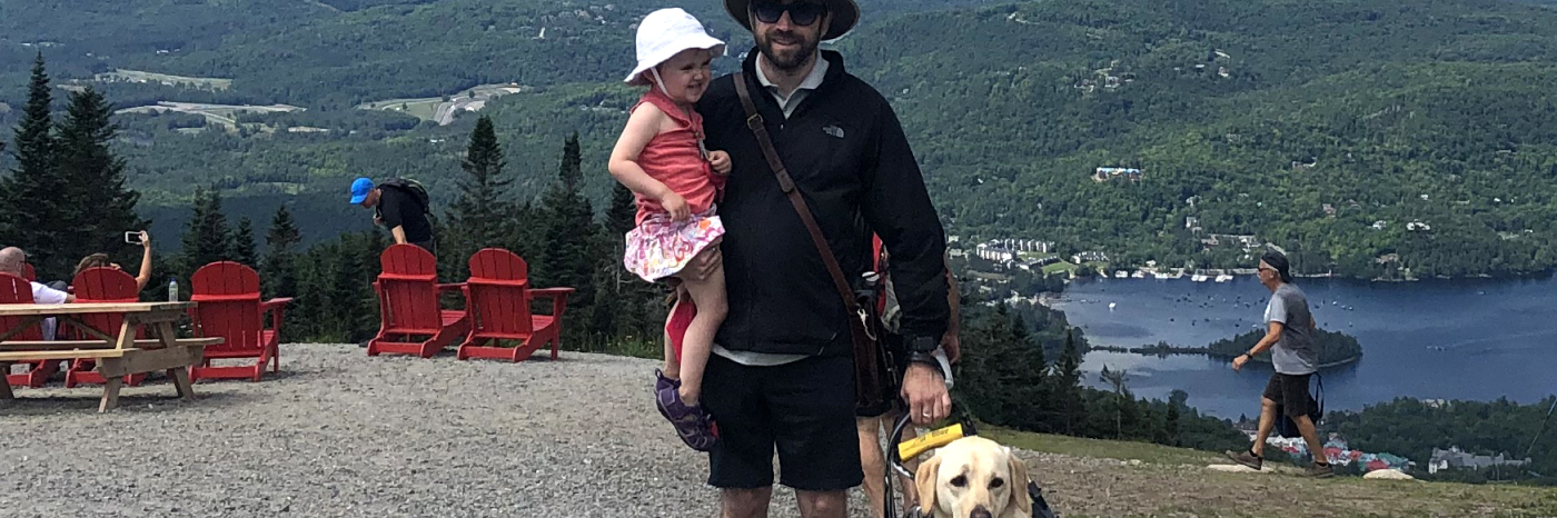 David with his daughter in one arm and his guide dog, Lilo's, harness in his other hand, smiling for the camera on Mont Tremblant overlooking Versant Sud, QB