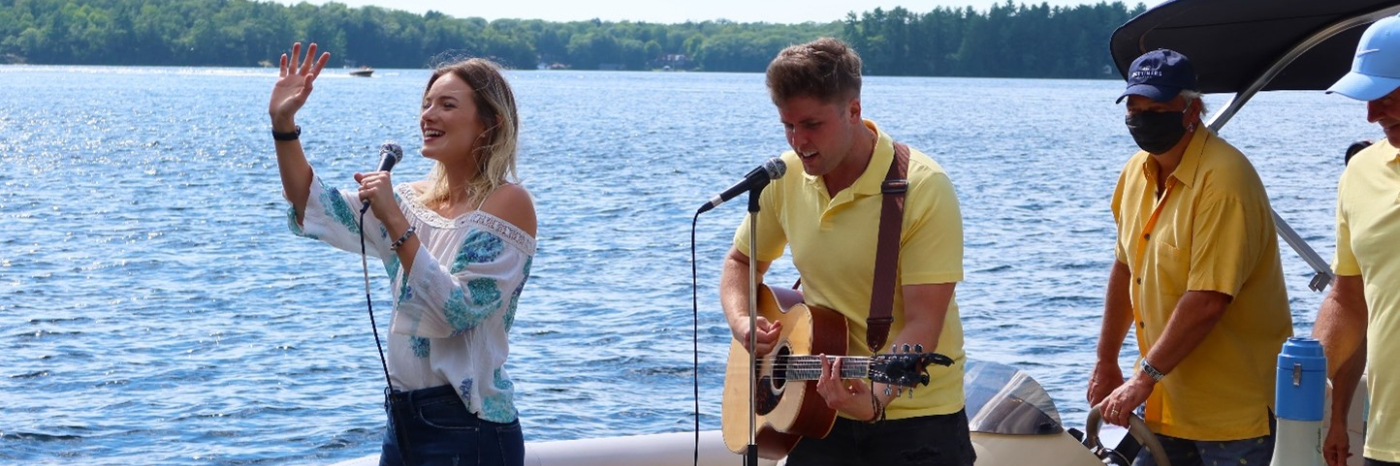 Leah Daniels singing and waving with Will Hebbes beside her playing guitar on a party pontoon, entertaining docks guests on Lake Muskoka.