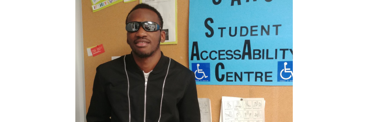  Eitel Houedakor smiling and standing in front of a bulletin board. He is wearing sunglasses and a hoodie.