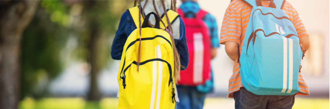 : The backs of three youth walking to school. They are wearing backpacks and are outdoors.