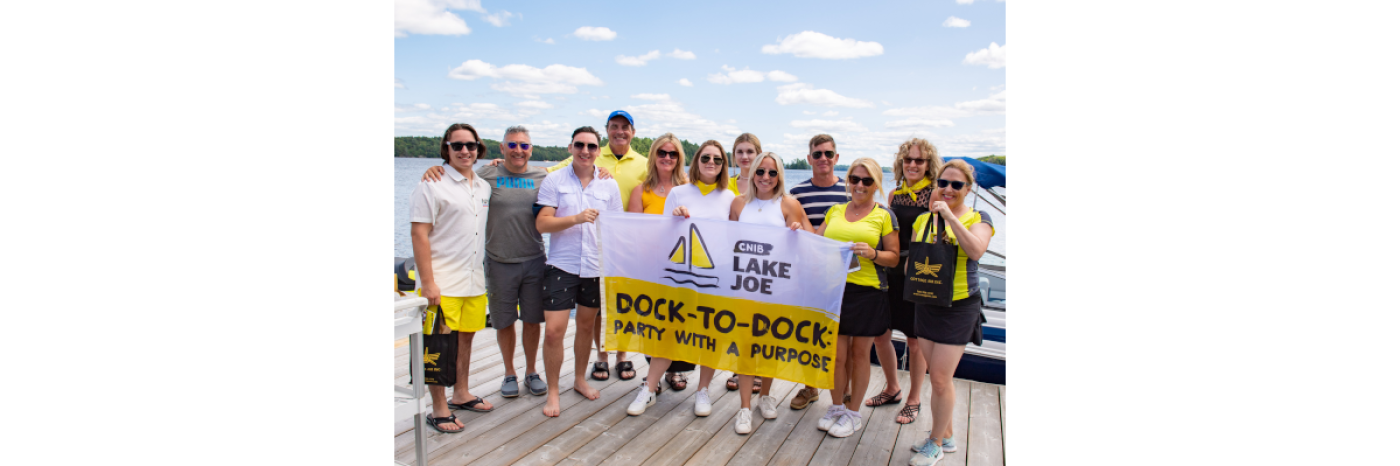 CNIB Muskoka Dock-to-Dock event volunteers posing at the CNIB Lake Joe boathouse, holding event flags.