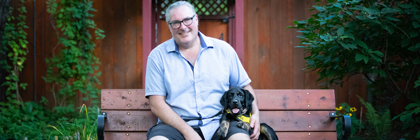 Christopher Adams sitting on a bench with a very young Percy sitting beside him wearing his bright yellow Future CNIB Guide Dogs vest.