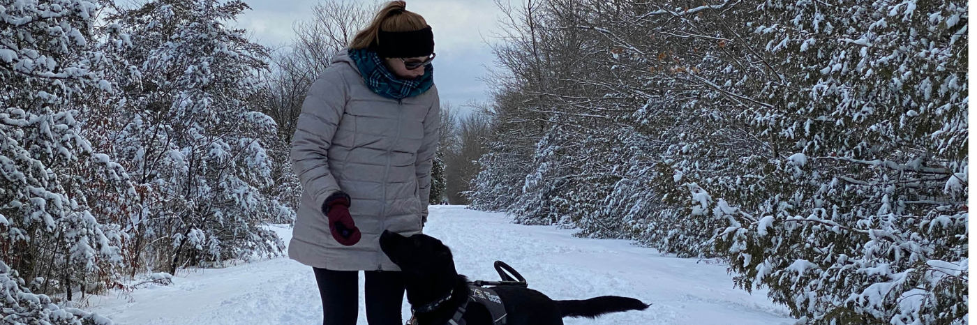 Kathleen, wearing a parka while walking down a snowy trail in the forest with her CNIB Guide Dog, Lily