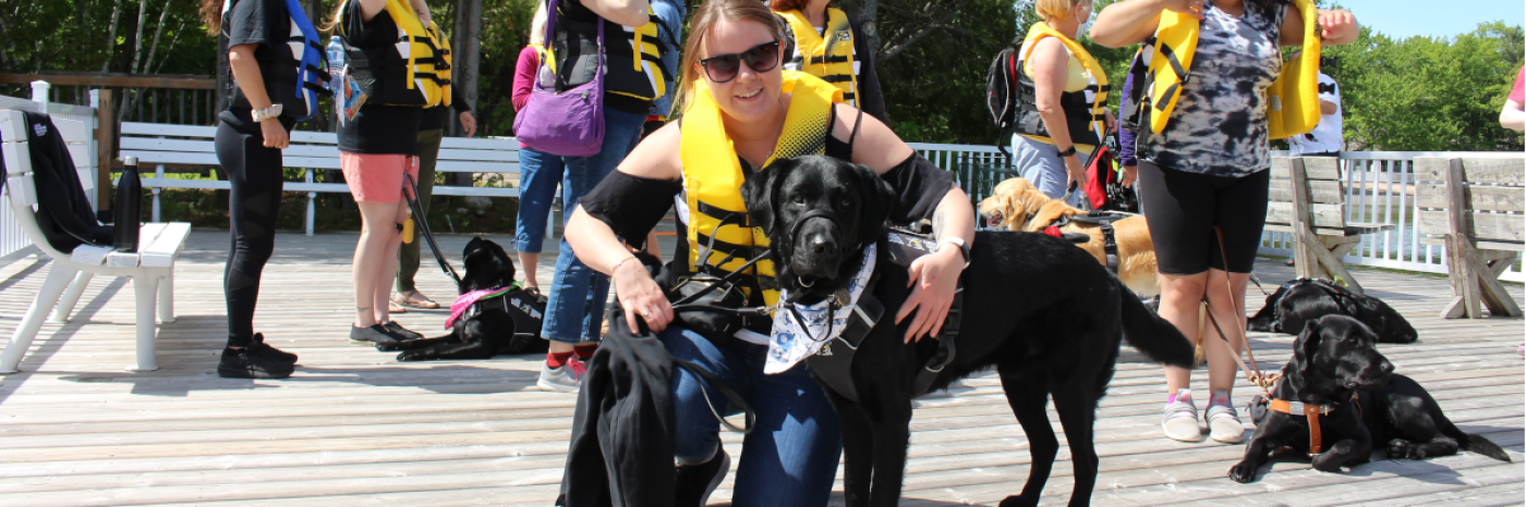  group of guide dog handlers wearing lifejackets stand on the dock at CNIB Lake Joe with their guide dogs. Taylor crouches and poses in the foreground next to her guide dog, Wallace.