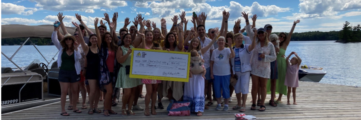 A group of guests stand on top of a waterfront dock and enthusiastically wave at the camera, showcasing their giant-cheque donation of $1,000 