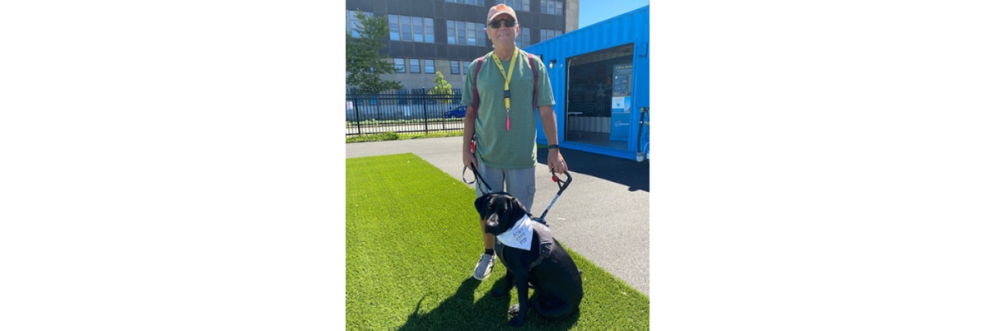 Blair and his black guide dog, Kelly, are outdoors standing on a patch of grass. Blair is holding Kelly's harness. 