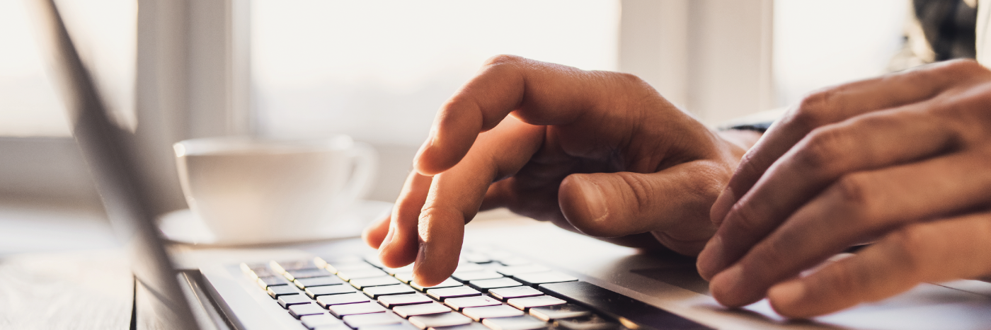 A close-up of a person’s hands typing on a laptop keyboard. The primary focus is on the hands and the keyboard, the background is blurred but it’s a well-lit indoor setting.
