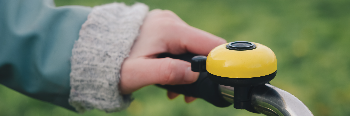 A person's hand rings a yellow bicycle bell. The bell is mounted on the bicycle handlebar.