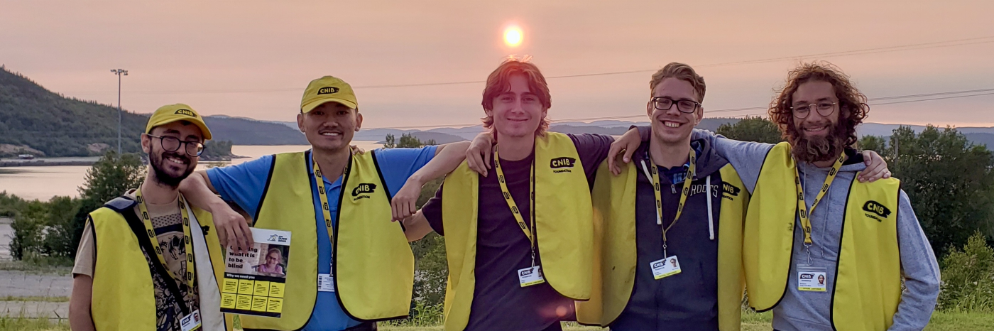 Five canvassers working on behalf of CNIB stand side-by-side with their arms around each other in front of a vibrant red sunset. All canvassers are wearing a yellow CNIB vest and lanyard with photo identification. The two canvassers on the left are also wearing yellow CNIB baseball caps and holding large cards that feature facts about blindness.