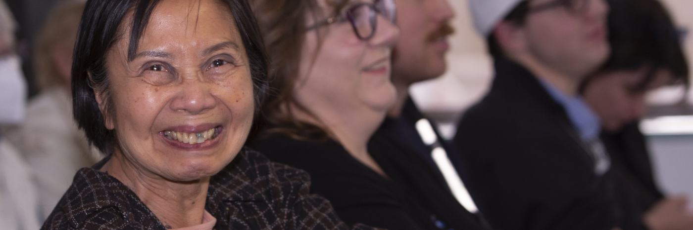 A woman with sight loss looks directly at the camera and smiles. She is sitting amongst a large group of people, who are out of focus in the background.