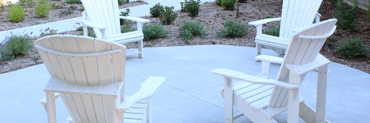 Four white Muskoka chairs are placed in a circle on a cement pad. There is a pathway through garden beds with small trees and shrubs.