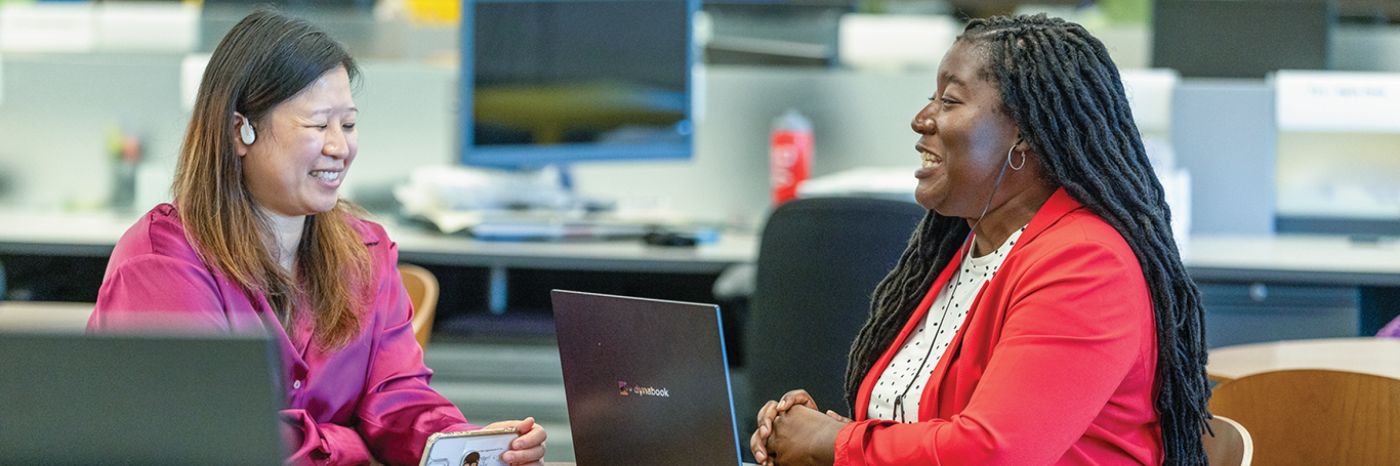 Two women sit at a table in an office environment, engaged in a conversation. The woman on the left is wearing a purple blouse. She is holding a smartphone in her hand and wears earbud headphones. The woman on the right has long braided hair and is wearing a red blazer. Both have laptops on the table in front of them. In the background are computers and different workstations.
