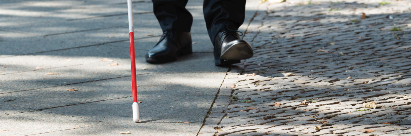 A white cane scans a sidewalk. In the background, men’s shoes trail behind the cane's tip.
