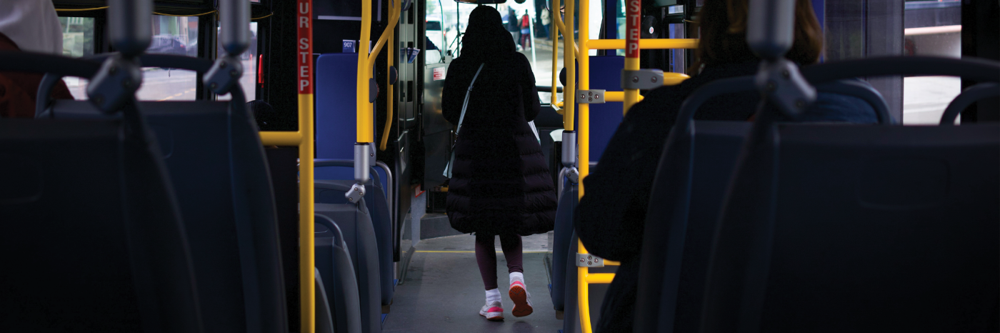 The interior of a city bus. The image is taken from the back of the bus, where a person walks down the aisle towards the front door.