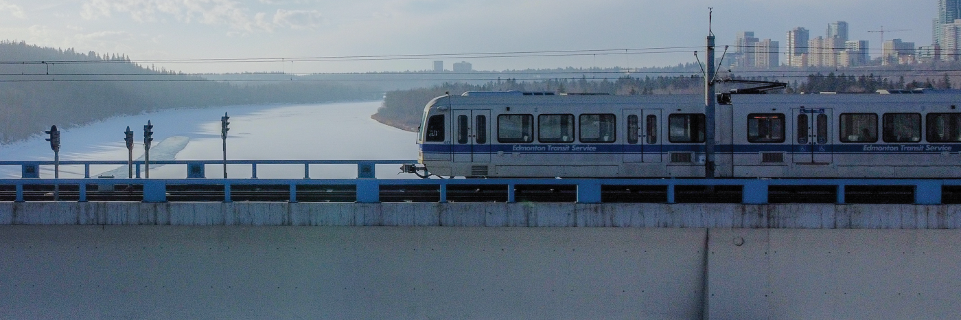 An Edmonton light-rail train travels across a bridge. The bridge is elevated above a frozen body of water, and the Edmonton skyline is in the background. 