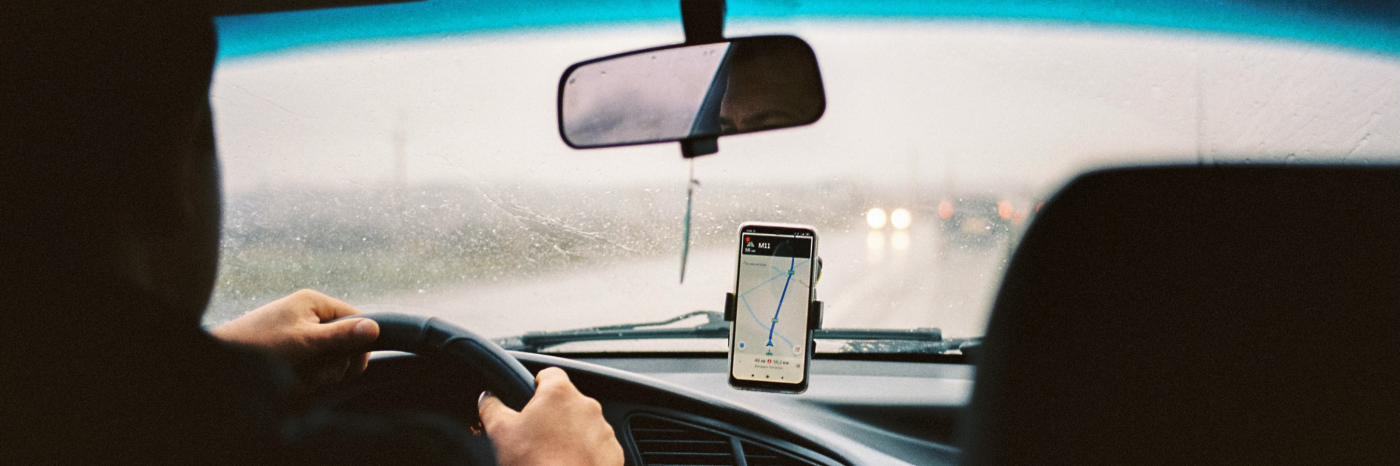 Inside a rideshare car, the photograph is taken from the passenger’s point of view in the back seat. There is a smartphone mounted on the dashboard of the car displaying a rideshare app. The driver’s hand is on the steering wheel and it is raining outside. 