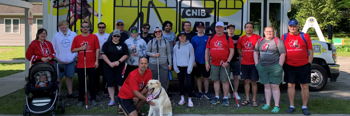 Members of the Canadian Blind Hockey Association (details) pose in front of the minibus at the CNIB Lake Joe Welcome Centre. They are wearing XX hockey jerseys and holding big hockey bags.  