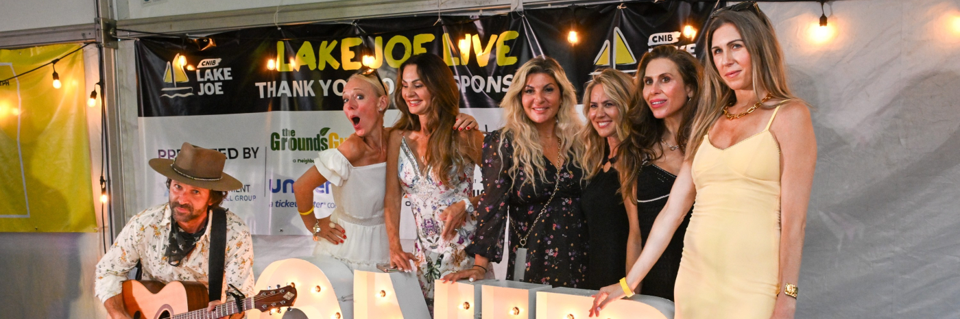 Six women wearing semi formal attire stand on a stage behind large lights in the shape of the letters C-N-I-B.