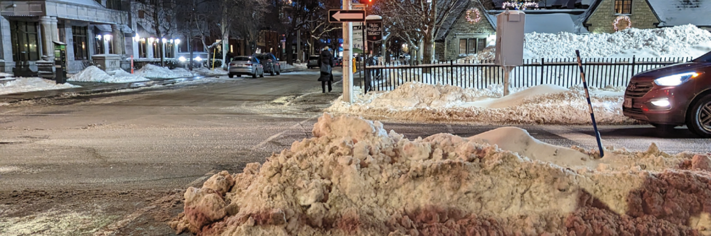 A pedestrian crosswalk in an urban neighbourhood is shown on a winter evening. The crosswalk is blocked by a snowbank piled several feet high.
