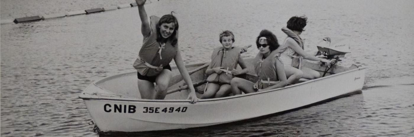 A black and white photo of two women and two children wearing lifejackets. They are smiling and sitting in a boat with CNIB on the side in the lake