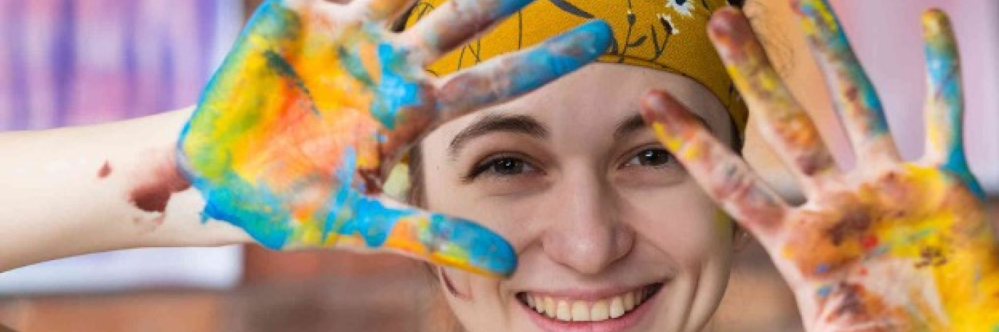 A young woman is smiling at the camera. Her hair is pulled back by a gold flowered bandana. She is holding her hands open in front of her, and they are covered in bright paint colours.