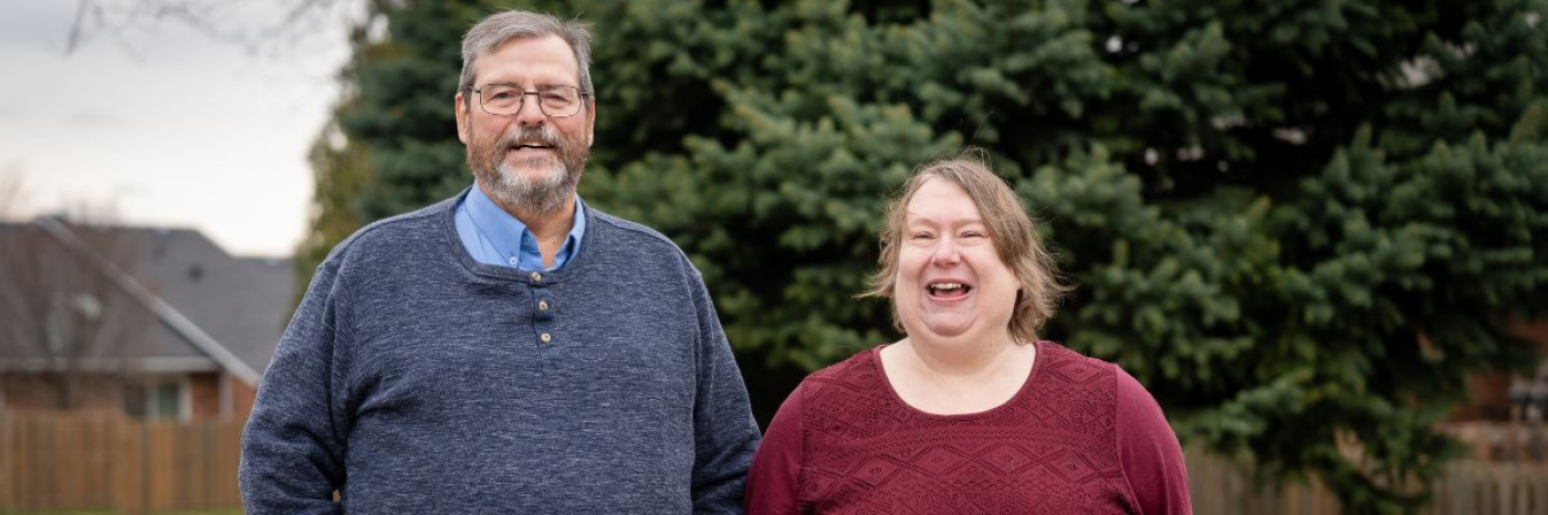 A man and woman stand together outdoors in front of a large evergreen tree, both smiling and laughing. The man, on the left, has grey hair, a beard, and glasses, and wears a navy blue henley sweater over a light blue shirt. The woman, on the right, has light brown hair and wears a dark burgundy textured top. The image is in a wide banner format.