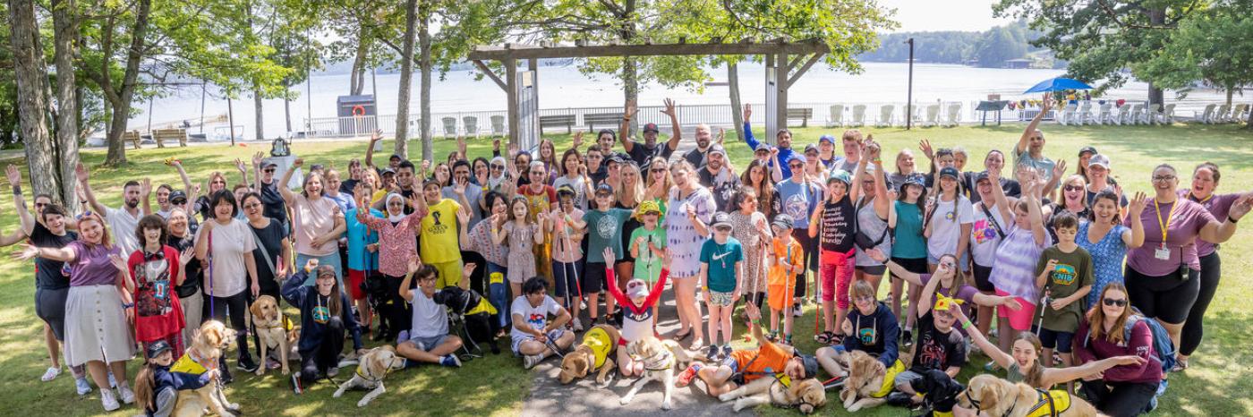 A large group of people of all ages, including many who are accompanied by buddy dogs or holding white canes, raise their hands in the air joyfully at Lake Joe - with the lake visible behind them.