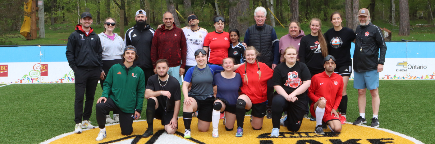 A group of blind soccer players of varying heights, ages, and skin tones stand in athletic gear on the accessible soccer pitch at CNIB Lake Joe, with a blind soccer ball in front of them.