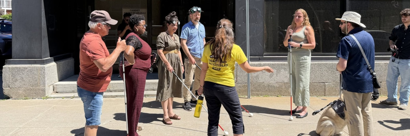 On a sidewalk, a woman wearing a yellow shirt is explaining something to a group of people. Among them are a man and a woman holding a white cane, a man accompanied by a guide dog, and two people wearing simulation goggles and a white cane.