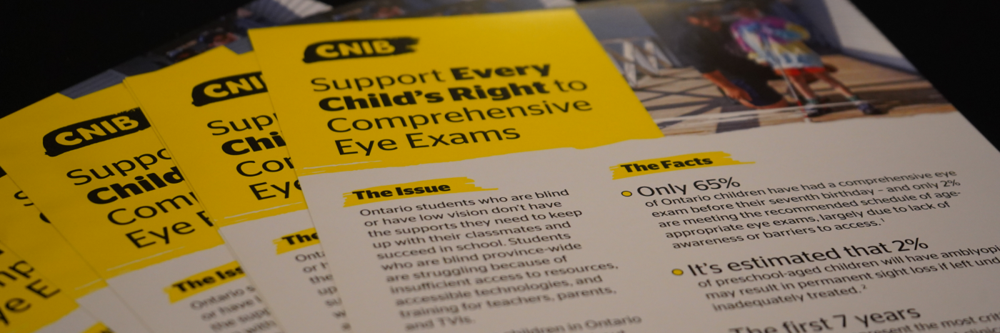 Several brochures are fanned out on a table. Each brochure has a yellow heading with a CNIB logo and black text that reads “Support Every Child’s Right to Comprehensive Eye Exams.” Subheadings are visible that read “The Issue” and “The Facts.”