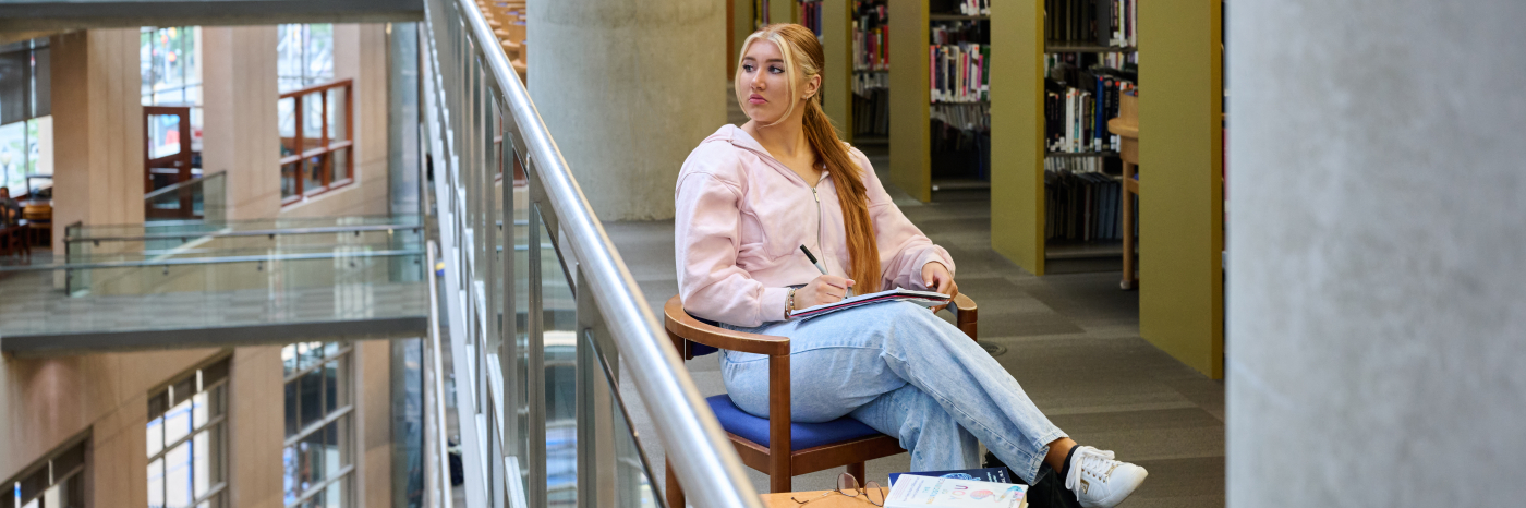 Ashlyn, wearing a pink sweater and jeans, sits in a wooden chair beside the glass railing of a multi floored library. She has a notebook and pen on her lap and is looking out over the railing's edge.