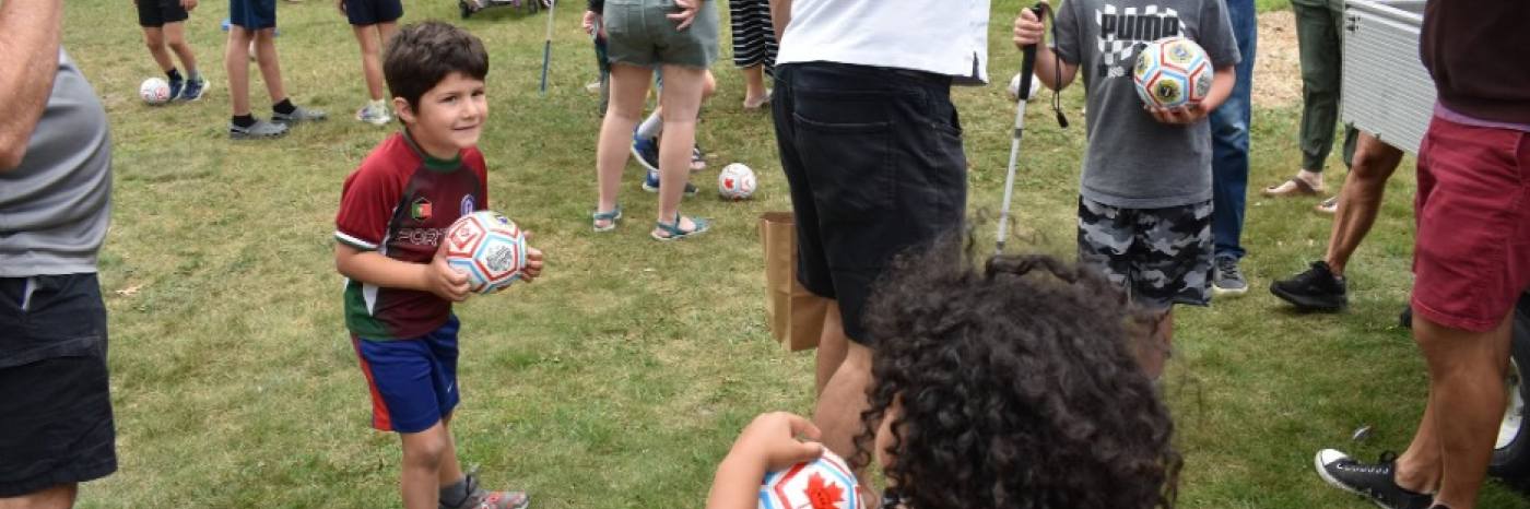 A crowd of children and adults gather on a grassy lawn for a soccer activity. Many of them are holding small white soccer balls in their hands or kicking them around their feet.