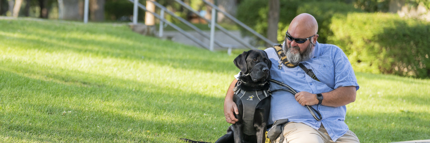 Jason sits on a park ledge beside Midas, his black guide dog. Jason is looking at Midas and smiling, and wears a light blue collared t-shirt , beige pants, and black sunglasses.