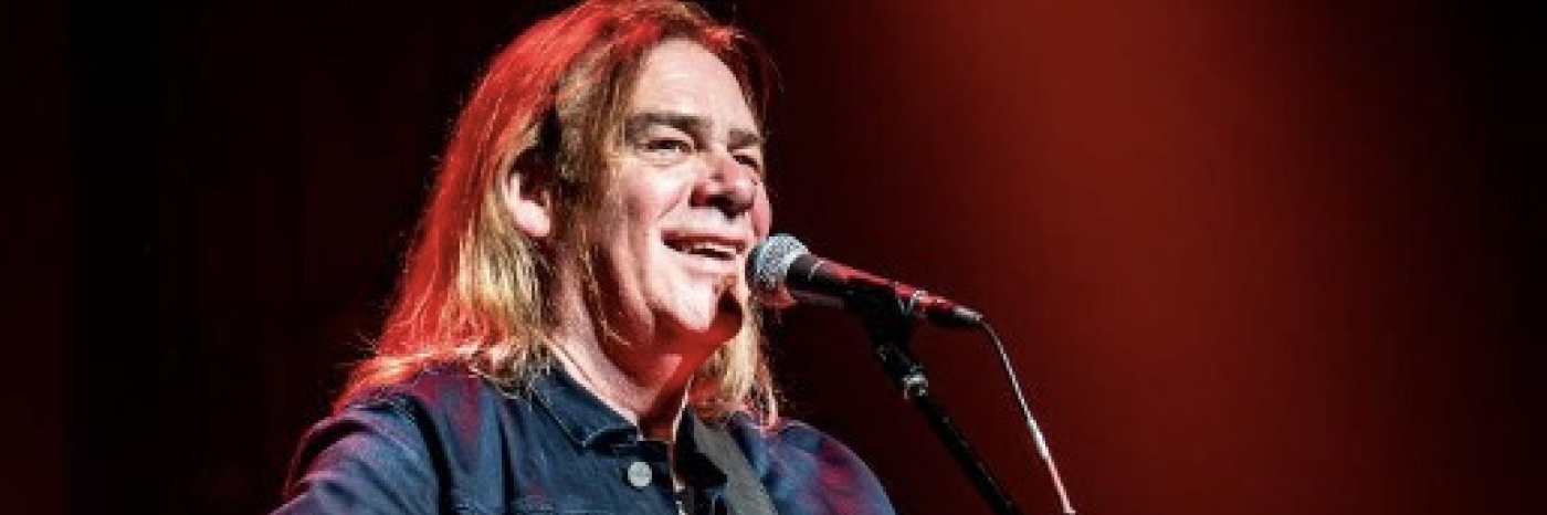 Alan Doyle smiles at the audience while performing onstage with an acoustic guitar at a microphone.