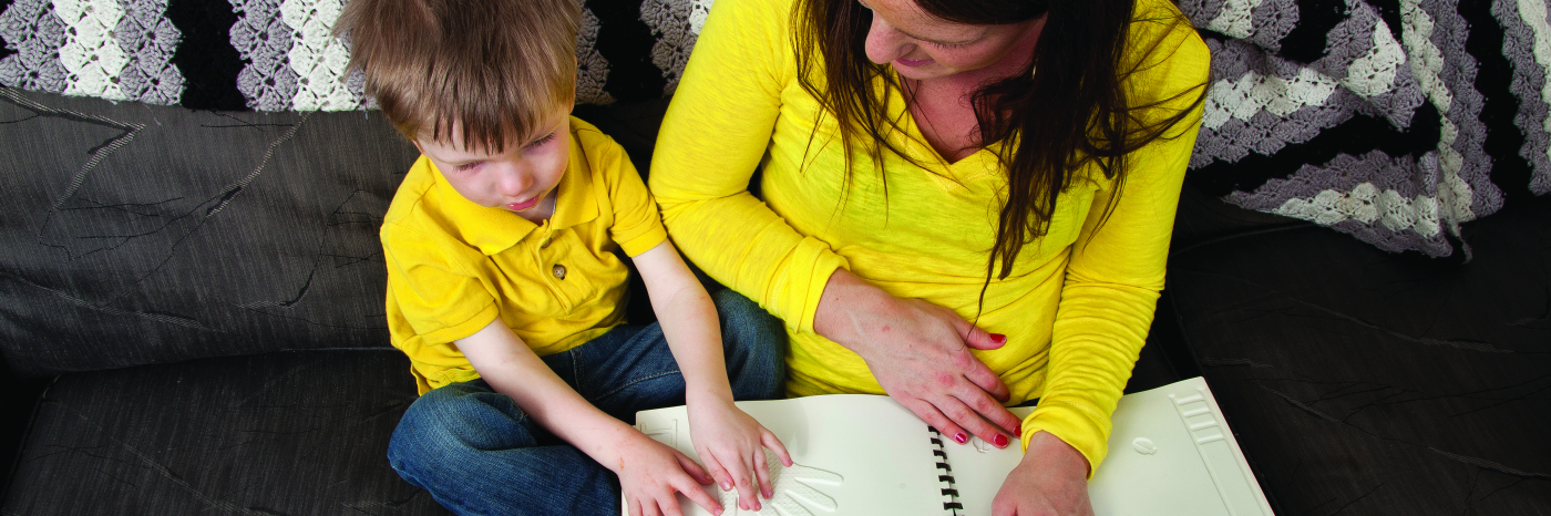 A little boy sits next to his mother on a couch reading a tactile book. Both are in yellow shirts.