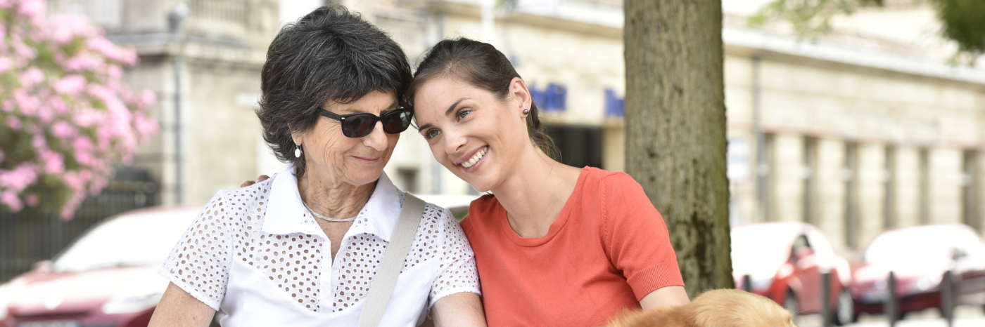 Two women sitting at a bench. The woman on the right has a white cane next to her, and the woman on the left has a golden labrador guide dog.