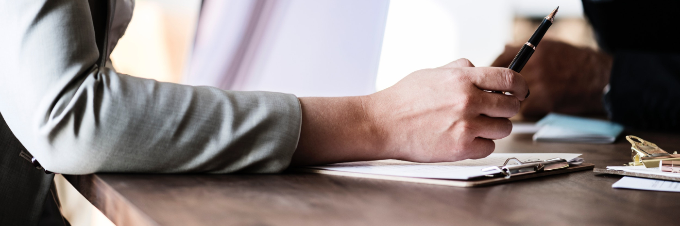 Close up of a person writing on a piece of paper with another person across the table from them