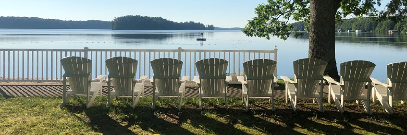 A row of white, Muskoka chairs sit on the shore of Lake Joe. 