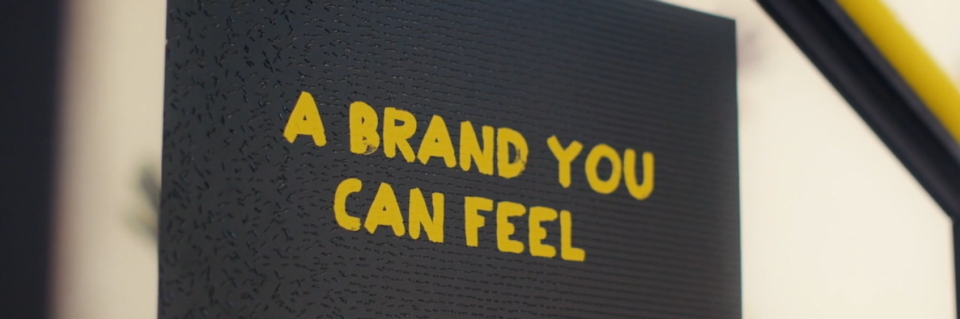 An image of a sign in braille with overlaid yellow CNIB branded letters in English reading A Brand You Can Feel