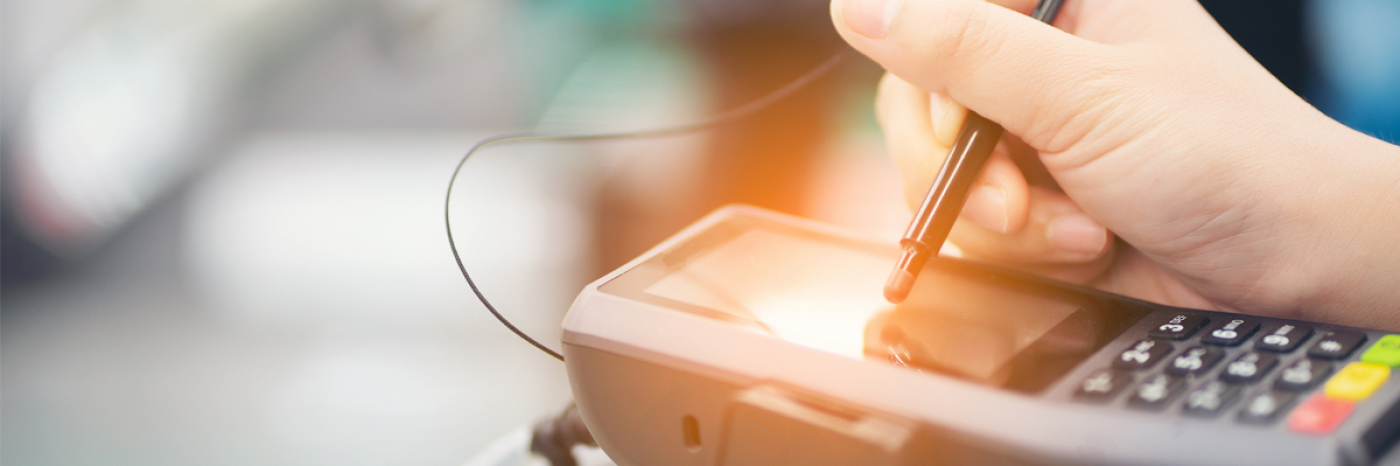 A hand using a stylus pen to make a selection on the touch screen surface of a payment terminal.