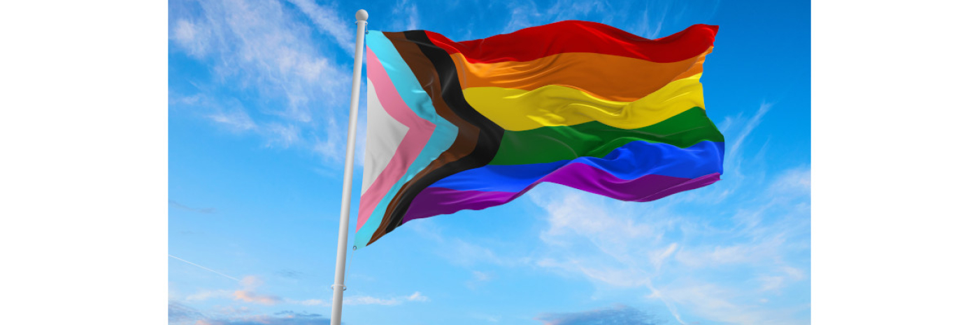 A Progress Pride flag waves in a cloudy sky.
