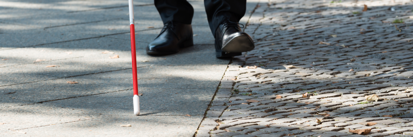 A white cane scans a sidewalk. In the background, mens shoes trail behind the tip of the cane.