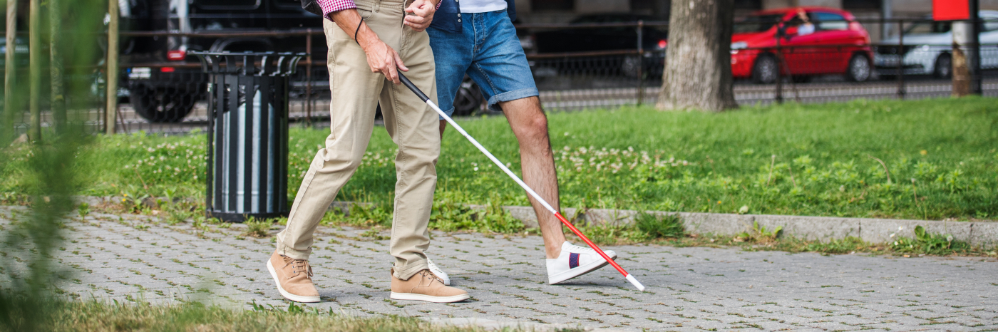 Two men take a walk along a paved path in a grassy area. One man navigates with a white cane.