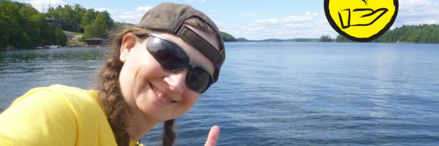 Jessica Bailey, poses at the Lake Joe waterfront. She does the American Sign Language sign for "I Love you". An icon of a hand with a heart floating above it appears in the top right hand corner. 