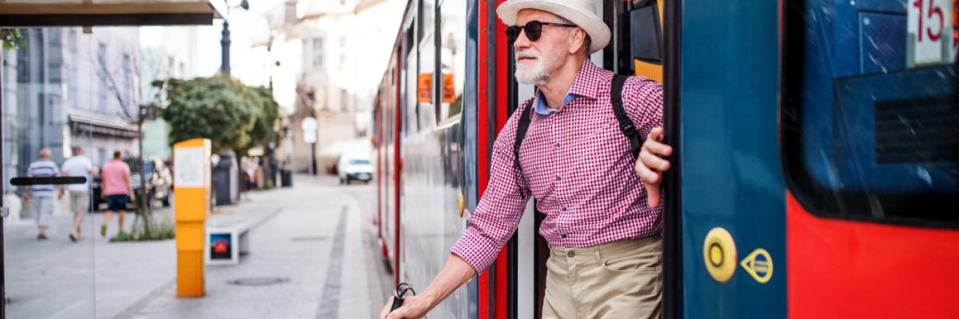 An image of an older man getting out of a streetcar and exiting onto a busy street. He uses a white cane, and is wearing a red and blue collared shirt, khakis and a fedora. He is smiling.